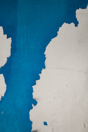 Peeling blue paint on a rough surface, revealing an underlying layer. The jagged lines of the chipped paint create an abstract and textured pattern, with stark contrast between the vibrant blue and the exposed lighter layer.