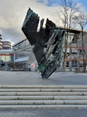 A large abstract sculpture made of dark, textured material stands on a paved area. The sculpture has jagged edges and appears modern and artistic. Behind it is a building with large glass windows and some graffiti on its walls. Several bicycles are parked nearby, and bare trees can be seen in the background.