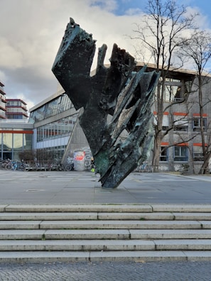 A large abstract sculpture made of dark, textured material stands on a paved area. The sculpture has jagged edges and appears modern and artistic. Behind it is a building with large glass windows and some graffiti on its walls. Several bicycles are parked nearby, and bare trees can be seen in the background.