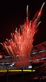 Brilliant red fireworks explode into the night sky over a stadium filled with spectators. The stadium is illuminated with colorful lights and digital displays showcasing text related to an event. The scene captures a festive and celebratory atmosphere.