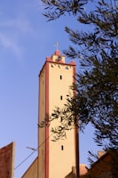 A tall beige tower with red accents stands prominently against a clear blue sky. The structure is topped with a cross and features small windows. Olive tree branches partially frame the tower, adding a touch of greenery to the scene.