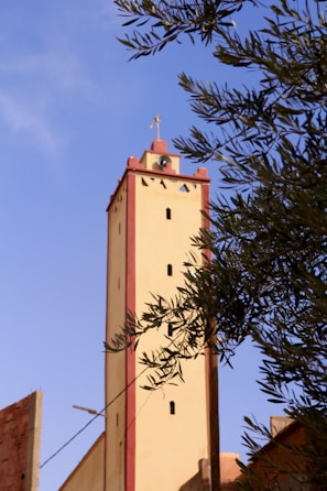 A tall beige tower with red accents stands prominently against a clear blue sky. The structure is topped with a cross and features small windows. Olive tree branches partially frame the tower, adding a touch of greenery to the scene.