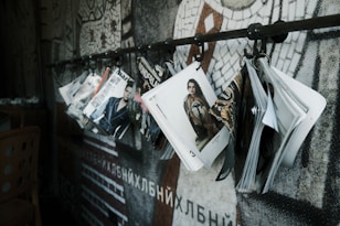 A series of fashion magazines hanging from hooks on a metal rod, with a focus on magazine covers that display models and fashion branding. The backdrop features a mosaic wall design with abstract patterns.