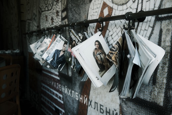 A series of fashion magazines hanging from hooks on a metal rod, with a focus on magazine covers that display models and fashion branding. The backdrop features a mosaic wall design with abstract patterns.