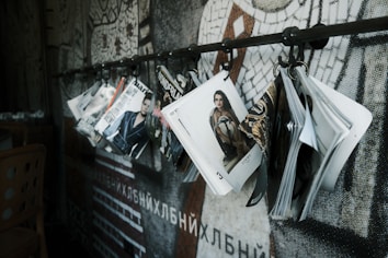 A series of fashion magazines hanging from hooks on a metal rod, with a focus on magazine covers that display models and fashion branding. The backdrop features a mosaic wall design with abstract patterns.
