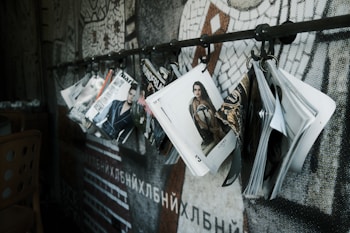 A series of fashion magazines hanging from hooks on a metal rod, with a focus on magazine covers that display models and fashion branding. The backdrop features a mosaic wall design with abstract patterns.
