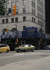 A bustling urban street with a vintage green car and a modern yellow taxi on the road. The scene is set in front of a historic bank building undergoing restoration. Traffic lights are visible, and there's a person walking on the sidewalk. The tall building has a classical architectural style with large windows and ornate details.