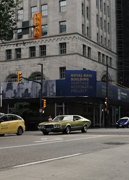 A bustling urban street with a vintage green car and a modern yellow taxi on the road. The scene is set in front of a historic bank building undergoing restoration. Traffic lights are visible, and there's a person walking on the sidewalk. The tall building has a classical architectural style with large windows and ornate details.