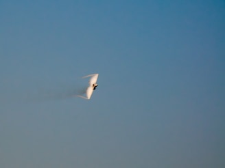 A jet aircraft is captured flying at high speed with a vapor cone forming around it, likely due to a rapid change in air pressure, against a clear blue sky.