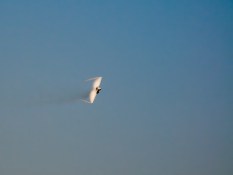 A jet aircraft is captured flying at high speed with a vapor cone forming around it, likely due to a rapid change in air pressure, against a clear blue sky.