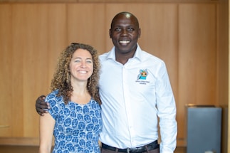 Two people are posing together indoors with a wooden wall background. The person on the left has curly hair and is wearing a blue patterned dress, while the person on the right is wearing a white shirt with the text 'Open Street Map Uganda'. Both are smiling and have a friendly demeanor.