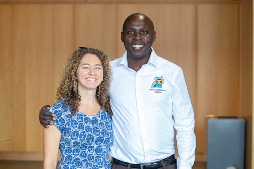 Two people are posing together indoors with a wooden wall background. The person on the left has curly hair and is wearing a blue patterned dress, while the person on the right is wearing a white shirt with the text 'Open Street Map Uganda'. Both are smiling and have a friendly demeanor.