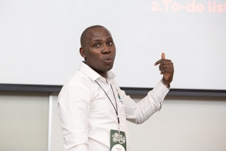 A person in a white shirt is standing in front of a blank screen or board, gesturing with one hand. They are wearing a name tag and appear to be making a point or giving a presentation.