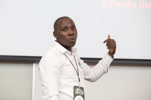 A person in a white shirt is standing in front of a blank screen or board, gesturing with one hand. They are wearing a name tag and appear to be making a point or giving a presentation.