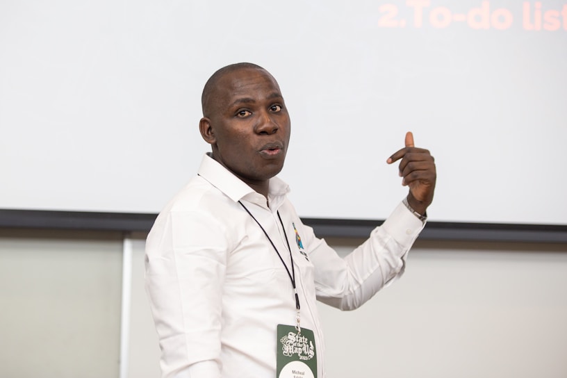 A person in a white shirt is standing in front of a blank screen or board, gesturing with one hand. They are wearing a name tag and appear to be making a point or giving a presentation.