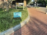 A sign with the message 'State of the Map US, Almost There!' is placed in a patch of grass next to a brick sidewalk. The scene is bathed in sunlight, casting shadows from nearby trees and lampposts. In the background, a building with a sign labeled 'VCU' is visible, alongside some greenery and a quiet street.