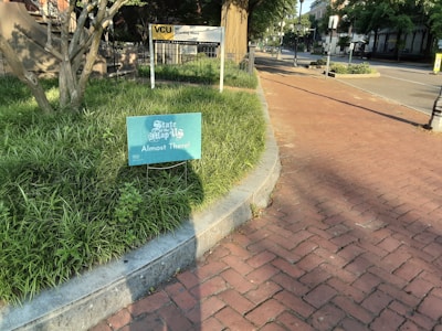A sign with the message 'State of the Map US, Almost There!' is placed in a patch of grass next to a brick sidewalk. The scene is bathed in sunlight, casting shadows from nearby trees and lampposts. In the background, a building with a sign labeled 'VCU' is visible, alongside some greenery and a quiet street.