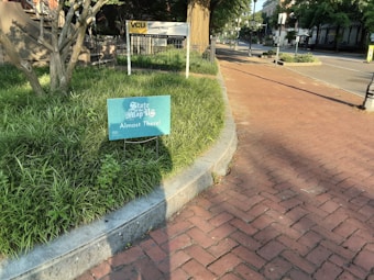 A sign with the message 'State of the Map US, Almost There!' is placed in a patch of grass next to a brick sidewalk. The scene is bathed in sunlight, casting shadows from nearby trees and lampposts. In the background, a building with a sign labeled 'VCU' is visible, alongside some greenery and a quiet street.