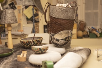A creative arrangement of handmade items on a table, including a basket, a lamp with a fur-covered shade, and various woven bowls. The items are made with a combination of woven materials and soft, furry textures. The background shows a cluttered workspace with various papers and tools.