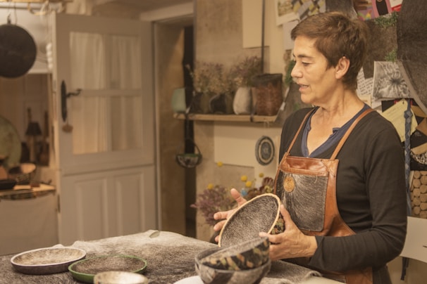 A person wearing a leather apron is standing in a workshop, holding handcrafted bowls. The room is filled with various materials and tools, with a door leading to another space. The walls have shelves with plants and decorative items, and papers pinned to a board suggest ongoing projects or inspiration.