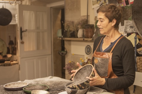 A person wearing a leather apron is standing in a workshop, holding handcrafted bowls. The room is filled with various materials and tools, with a door leading to another space. The walls have shelves with plants and decorative items, and papers pinned to a board suggest ongoing projects or inspiration.