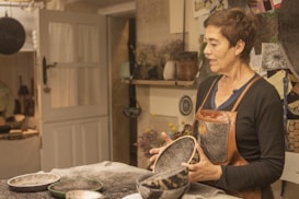 A person wearing a leather apron is standing in a workshop, holding handcrafted bowls. The room is filled with various materials and tools, with a door leading to another space. The walls have shelves with plants and decorative items, and papers pinned to a board suggest ongoing projects or inspiration.