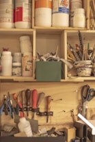 A well-organized workshop wall with various tools and materials. The shelves contain jars and containers filled with supplies like paint and other materials. Below the shelves, there are hand tools including screwdrivers, pliers, and carving tools hanging on the wall. A pair of scissors is visible on the right side, and a basket holds pens and brushes.