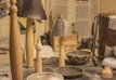 A collection of handcrafted items on a table including wooden candle holders, lamps with gray textured shades, and decorative bowls. A woven leather bag is visible on the right, while various materials and tools can be seen in the background.