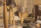 A collection of handcrafted items on a table including wooden candle holders, lamps with gray textured shades, and decorative bowls. A woven leather bag is visible on the right, while various materials and tools can be seen in the background.