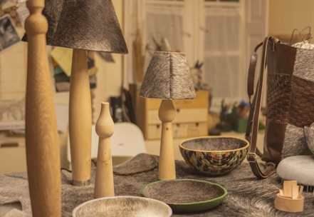 A collection of handcrafted items on a table including wooden candle holders, lamps with gray textured shades, and decorative bowls. A woven leather bag is visible on the right, while various materials and tools can be seen in the background.