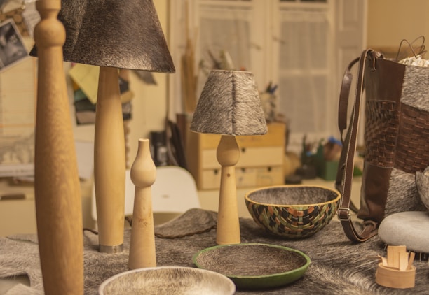 A collection of handcrafted items on a table including wooden candle holders, lamps with gray textured shades, and decorative bowls. A woven leather bag is visible on the right, while various materials and tools can be seen in the background.