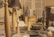 A collection of handcrafted items on a table including wooden candle holders, lamps with gray textured shades, and decorative bowls. A woven leather bag is visible on the right, while various materials and tools can be seen in the background.