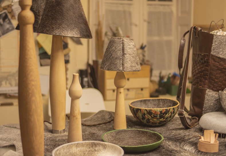 A collection of handcrafted items on a table including wooden candle holders, lamps with gray textured shades, and decorative bowls. A woven leather bag is visible on the right, while various materials and tools can be seen in the background.