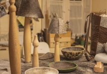 A collection of handcrafted items on a table including wooden candle holders, lamps with gray textured shades, and decorative bowls. A woven leather bag is visible on the right, while various materials and tools can be seen in the background.