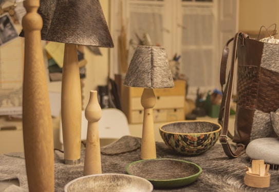A collection of handcrafted items on a table including wooden candle holders, lamps with gray textured shades, and decorative bowls. A woven leather bag is visible on the right, while various materials and tools can be seen in the background.