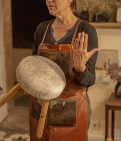 A person is wearing a brown leather apron and holding a drum-like instrument with a round, furry surface and wooden handles. The setting seems to be indoors with warm lighting, and there are various items on a shelf in the background.