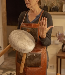 A person is wearing a brown leather apron and holding a drum-like instrument with a round, furry surface and wooden handles. The setting seems to be indoors with warm lighting, and there are various items on a shelf in the background.