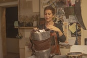 A person stands in a rustic room, focused on crafting a handmade item. They are holding a woven item made of natural materials, possibly a basket. The room has a cozy feel with several craft-related materials and tools around. There are shelves with various items and a bulletin board with notes and images in the background. The person is wearing an apron, suggesting they are engaged in some form of artisanal work.