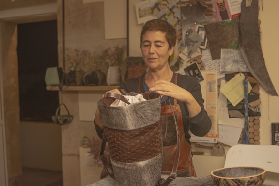 A person stands in a rustic room, focused on crafting a handmade item. They are holding a woven item made of natural materials, possibly a basket. The room has a cozy feel with several craft-related materials and tools around. There are shelves with various items and a bulletin board with notes and images in the background. The person is wearing an apron, suggesting they are engaged in some form of artisanal work.