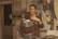 A person stands in a rustic room, focused on crafting a handmade item. They are holding a woven item made of natural materials, possibly a basket. The room has a cozy feel with several craft-related materials and tools around. There are shelves with various items and a bulletin board with notes and images in the background. The person is wearing an apron, suggesting they are engaged in some form of artisanal work.