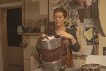 A person stands in a rustic room, focused on crafting a handmade item. They are holding a woven item made of natural materials, possibly a basket. The room has a cozy feel with several craft-related materials and tools around. There are shelves with various items and a bulletin board with notes and images in the background. The person is wearing an apron, suggesting they are engaged in some form of artisanal work.