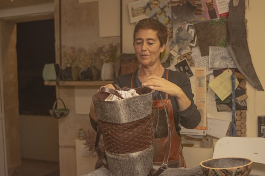 A person stands in a rustic room, focused on crafting a handmade item. They are holding a woven item made of natural materials, possibly a basket. The room has a cozy feel with several craft-related materials and tools around. There are shelves with various items and a bulletin board with notes and images in the background. The person is wearing an apron, suggesting they are engaged in some form of artisanal work.