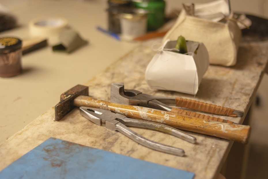 A wooden workbench with various tools including pliers, a hammer, and a chisel. There are also containers and bags in the background, suggesting a workshop or crafting environment.
