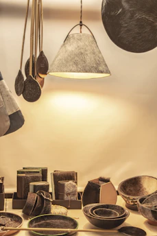 A collection of handcrafted items sits on display. The setup includes a variety of objects such as bowls, trays, and a hanging lamp, all of which have a fur or leather-like texture. The scene is warmly lit, enhancing the natural materials of each item. Wooden utensils with a dark finish are suspended next to the lamp.