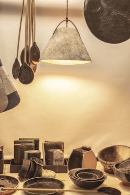 A collection of handcrafted items sits on display. The setup includes a variety of objects such as bowls, trays, and a hanging lamp, all of which have a fur or leather-like texture. The scene is warmly lit, enhancing the natural materials of each item. Wooden utensils with a dark finish are suspended next to the lamp.