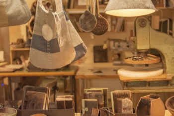 A cozy workshop setting with various leather goods and accessories on display. Handcrafted items such as wallets, bags, and keychains are neatly arranged on a wooden table. There is a sewing machine in the background, along with hanging lamps that provide warm lighting. The ambiance suggests a focus on craftsmanship and artisanal work.