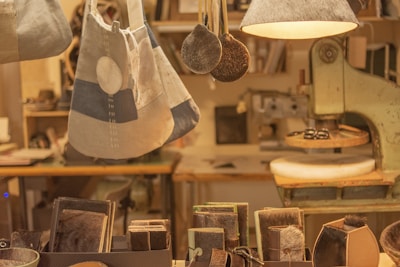 A cozy workshop setting with various leather goods and accessories on display. Handcrafted items such as wallets, bags, and keychains are neatly arranged on a wooden table. There is a sewing machine in the background, along with hanging lamps that provide warm lighting. The ambiance suggests a focus on craftsmanship and artisanal work.