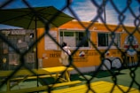 A person is standing at a bright yellow food truck, which has a menu and large text advertising fresh fries and cold drinks. The scene is viewed through a chain-link fence. A yellow picnic bench and a large umbrella can be seen in front of the food truck. The sky is clear blue with some wispy clouds.