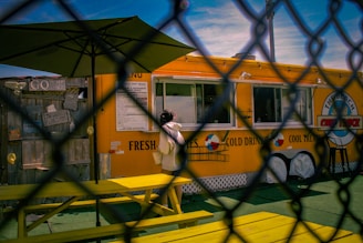 A person is standing at a bright yellow food truck, which has a menu and large text advertising fresh fries and cold drinks. The scene is viewed through a chain-link fence. A yellow picnic bench and a large umbrella can be seen in front of the food truck. The sky is clear blue with some wispy clouds.