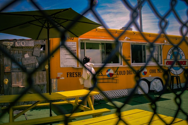A person is standing at a bright yellow food truck, which has a menu and large text advertising fresh fries and cold drinks. The scene is viewed through a chain-link fence. A yellow picnic bench and a large umbrella can be seen in front of the food truck. The sky is clear blue with some wispy clouds.